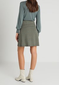 Haut vert à manches longues associé à une jupe patineuse imprimée, de longueur genoux, arborant des motifs géométriques dans des tons terreux et des bottines beiges.