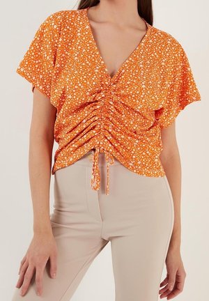 Frau trägt eine kurzärmelige orange geblümte, geraffte Bluse mit V-Ausschnitt und beige eng anliegende Hose, gezeigt vom Hals bis zu den Hüften.