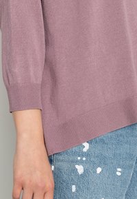 Heller lila gestrickter Pullover mit geripptem Saum und kurzen Ärmeln, kombiniert mit hellen Jeans mit Farbspritzern.