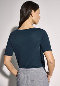 Tílko s kulatým výstřihem a krátkým rukávem v tmavě tyrkysové bavlně, mírně přiléhavé v ramenních oblastech, spárováno se světle šedými kalhotami s elastickým pasem a kapsami.