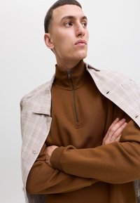 Fiatal férfi rövid hajjal, barna cipzáras pulóvert viselve és világos bézs kockás dzsekit a vállán átvetve, karjait keresztezi.