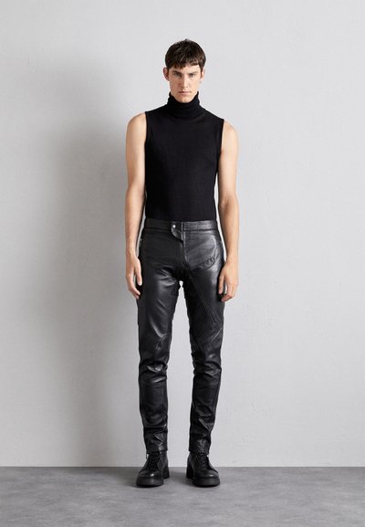Črna brezrokavna turtleneck majica v kombinaciji s tesnimi črnimi usnjeni hlačami z angularnimi šivi in zadrgami na žepih. Črne čevlje z vezalkami dopolnjujejo videz.