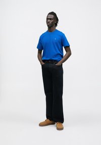 Niebieski t-shirt z okrągłym dekoltem, czarne jeansy o prostych nogawkach i brązowe trampki. Model stoi z rękami w kieszeniach na jednolitym białym tle.