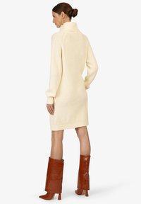 Crème ribbel turtleneck jurk valt tot halverwege de dij, gecombineerd met glanzende bruine krokodillen-embossed knielaarzen met blokhakken.