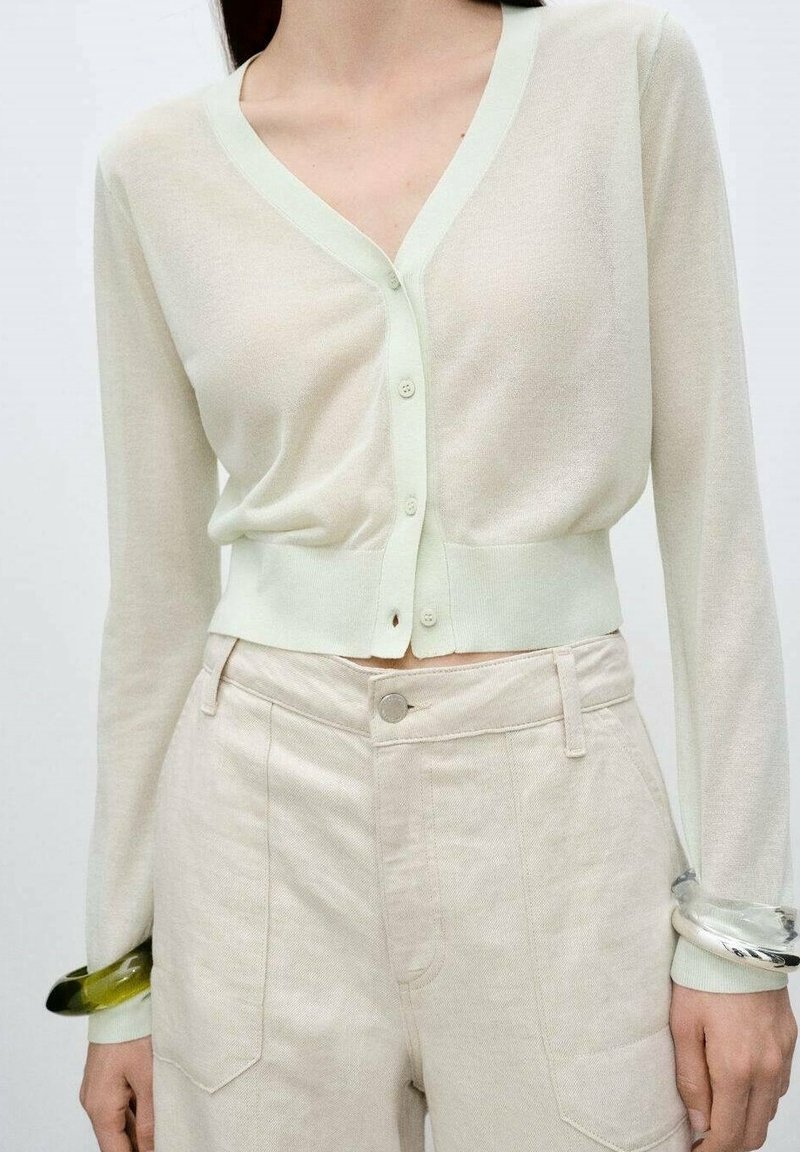 Femme portant un cardigan court beige à col en V avec boutons et un pantalon beige clair taille haute, avec des bracelets transparents aux poignets.