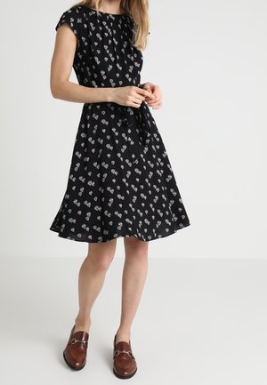 Femme portant une robe noire mi-longue avec un motif de montgolfières et des mocassins marron, debout devant un fond clair uni.