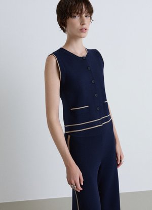 Donna con capelli corti che indossa un top senza maniche blu navy abbottonato e pantaloni abbinati con rifiniture beige, in piedi davanti a uno sfondo chiaro e uniforme.