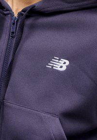 Μπλε ναυτικό φούτερ με φερμουάρ, με λευκό λογότυπο New Balance κεντημένο στο στήθος και ορατή ραφή εμπρός στις τσέπες.