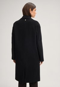 Zwarte wollen jas met rechte snit, notch-kraag en een enkele rugventilatie. Bevat een klein rond knopdetail bij de hals.