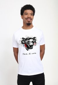 T-shirt bianco in cotone con un'illustrazione di testa di mucca in nero e grigio, dettagli rossi e blue, con la scritta "Tâche de vache" stampata sotto.