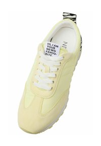 Yellow sneaker with suede and textile upper, white laces, rubber sole, and a black-and-white striped heel tab. Tag with text on the tongue.