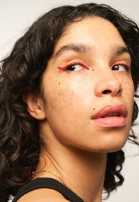 Primo piano del viso che mostra capelli neri ricci di lunghezza media. Le caratteristiche notevoli includono un design di eyeliner rosso, lentiggini e un colore naturale delle labbra.