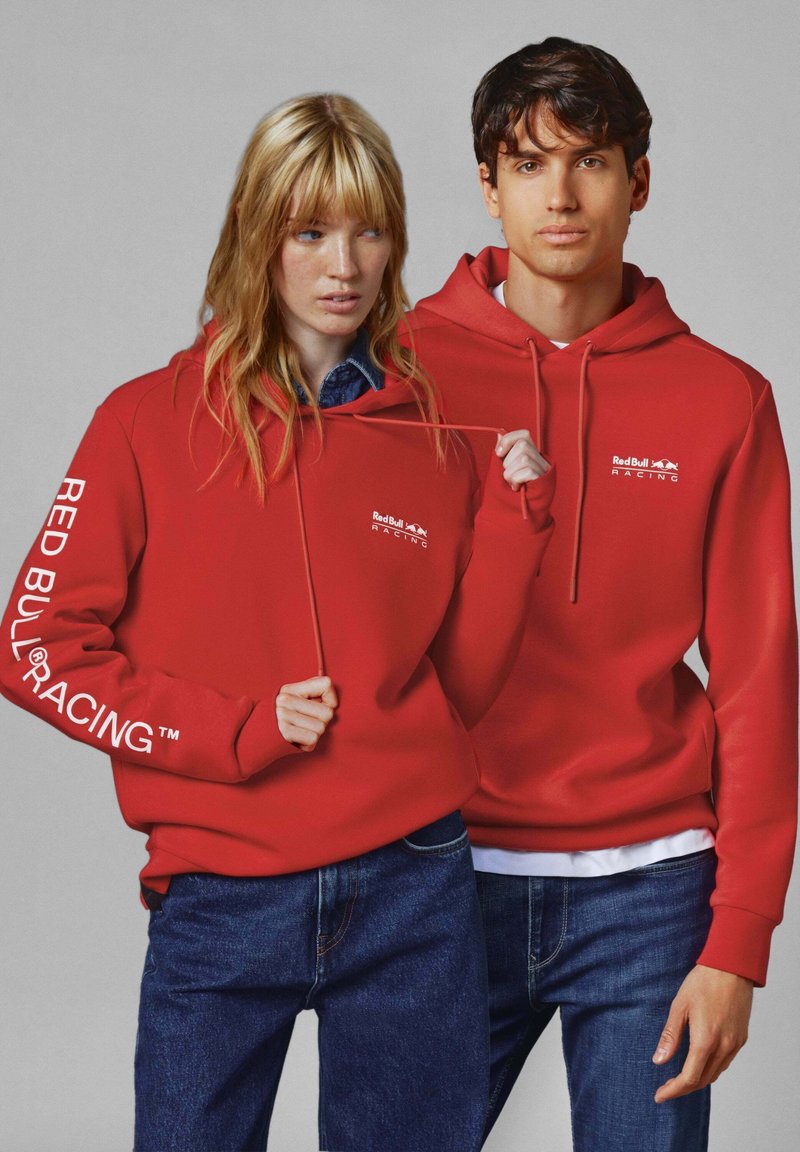 Rdeče kapuce s vrvico, ki imajo logotip "Red Bull Racing" in belo besedilo na eni rokavu, v kombinaciji s temno denim jeansom.