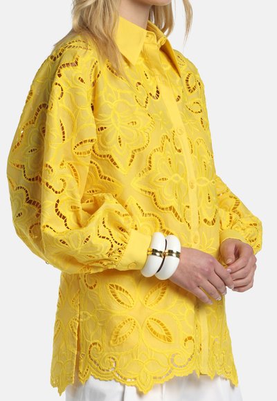 Sieviete valkā spilgti dzeltenu mežģīņu blūzi ar ziedu rakstiem un pūstām piedurknēm, aksesuāros divas masīvas baltas aproces.