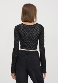 Top corto in pizzo nero con maniche lunghe, caratterizzato da un motivo floreale. Indossato con pantaloni in denim neri a vita alta, che mettono in risalto il design aderente.