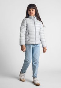 Ljust grå puffjacka med horisontell quiltad design, stående krage och dragkedjefickor. Bärs över blå jeans och vita sneakers.
