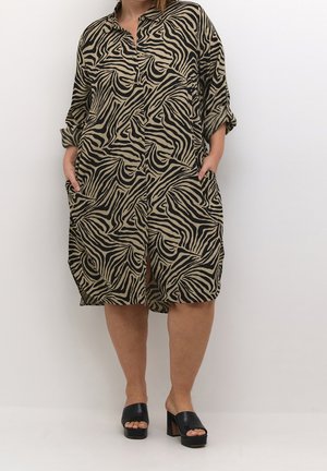 Kvinde iført knælang beige og sort zebraprint skjortekjole med opsmøgede ærmer og sorte åbne blockhælsandaler, hænderne i lommerne.