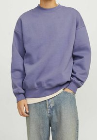 Person, der einen schlichten lila Rundhals-Sweatshirt über einem beigen Hemd und hellblauen Jeans trägt, mit einer Hand in der Tasche und einer entspannt.
