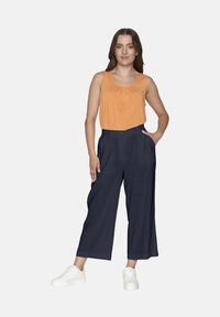 Orange ärmlös topp med en rynkad krage, parat med marinblå vida byxor med sidofickor. Bärs med vita sneakers.