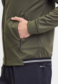 Giacca zip-up verde oliva con tasche laterali e vita nera caratterizzata da due strisce bianche. Texture liscia e design a vestibilità atletica.