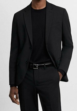 Hombre con chaqueta de traje negra, camisa negra, pantalones negros y cinturón negro con hebilla plateada, mano en el bolsillo.