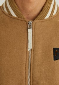 Brun ull bomberjacka med randig ribbad krage, silverfärgad dragkedja och en liten svart lapp med orange accent.