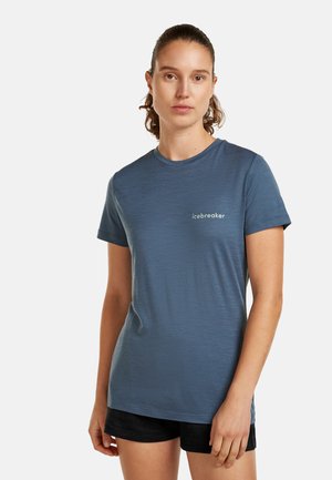 Jeune femme aux cheveux bruns attachés en queue de cheval, portant un t-shirt bleu à manches courtes Icebreaker et un short noir, debout devant un fond clair uni.