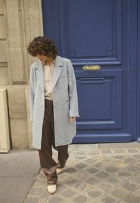 Cappotto azzurro a motivo houndstooth sopra una camicia beige, pantaloni larghi marroni e scarpe beige a punta; in piedi su una strada in ciottoli.