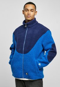 Blue and navy fleece jacket featuring a high collar, zip closure, and contrasting color panels. Textured, warm material with logo detail.
