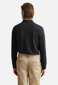 Hombre de espaldas con una camisa negra de manga larga metida en unos pantalones beige con bolsillos traseros con botones, sobre un fondo liso.