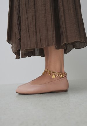 Voeten met lichtroze ballerina’s en een gouden enkelbandje met bedel, met een bruine geplooide rok zichtbaar boven de enkels, staande op een grijze vloer.