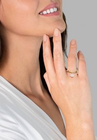 Auksinis žiedas su mažyte širdel�ės formos deimanto pakabuku, dėvimas ant rankos, kuri liesia smakrą. Odos tonas šviesus; fonas neutralus.
