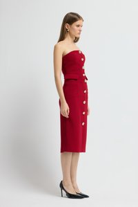 Rotes trägerloses Kleid mit tailliertem Oberteil und Bleistiftrock. Verziert mit goldenen Knöpfen und seitlichen Taschen. Kombiniert mit schwarzen Stiletto-Absätzen.