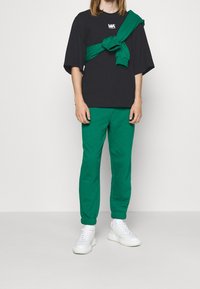 Person iført sort oversized t-shirt med hvidt logo, grøn sweatshirt bundet om brystet, matchende grøn joggingbukser og hvide sneakers.
