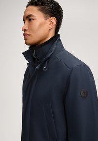 Marineblauwe buitenjas met een hoge kraag, knopen- en ritsafsluitingen, gestructureerde stof en een logopatch op de mouw.