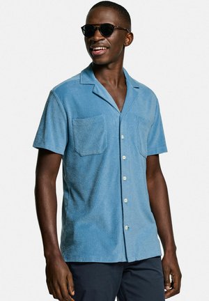 Hellblaues kurzärmliges Hemd aus strukturiertem Stoff, mit Kragen, zwei aufgesetzten Taschen und weißen Knöpfen. Getragen mit Sonnenbrille.