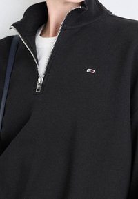 Felpa nera con zip realizzata in un mix di cotone. Presenta un colletto alto, una zip argentata e un piccolo logo ricamato in rosso, bianco e blu.