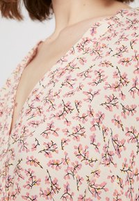 Kukkakuosisella kankaalla, jossa on pieniä vaaleanpunaisia ja keltaisia kukkia vaaleankreemivalkoisella taustalla. Kangas on kevyesti strukturoidulla ja kevyellä designilla sekä V-aukko.