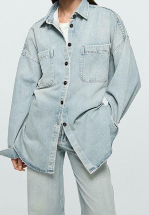 Personne portant une veste en jean oversize bleu clair avec de grandes poches avant et un pantalon en jean large assorti.
