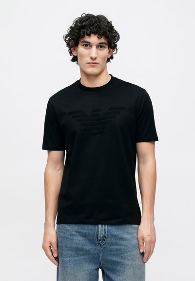 Μαύρο βαμβακερό μπλουζάκι με υφασμένο λογότυπο αετού στο κέντρο, στρογγυλή λαιμόκοψη, κοντά μανίκια και άνετη εφαρμογή, σε συνδυασμό με ανοιχτό μπλε τζιν.