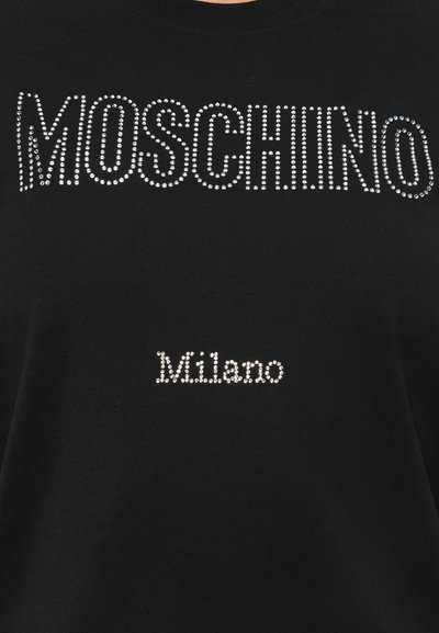 Melns T-krekls ar uzrakstu "MOSCHINO" lieliem rhinestones augšpusē un "Milano" mazākiem rhinestones zemāk. Īsas piedurknes un apaļa kakla izgriezums.