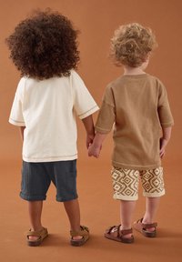 Deux enfants se tiennent la main, portant une chemise crème clair et une chemise marron, des shorts cargo gris foncé et des shorts beiges à motifs avec des sandales marron.