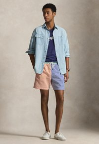 Νεαρός άνδρας που φοράει γαλάζιο πουκάμισο με κουμπιά πάνω από μπλε μπλουζάκι, πολύχρωμα κοντά σορτς καρό και λευκά αθλητικά παπούτσια, στέκεται μπροστά σε ουδέτερο φόντο.