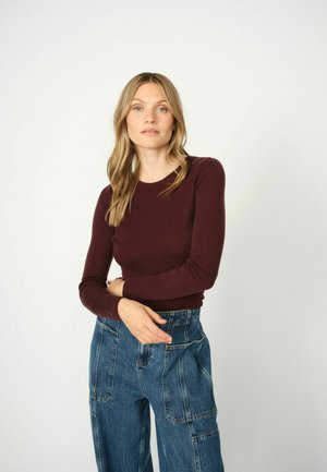 Burgunderfarbene Langarmbluse, taillierte Passform, kombiniert mit hochgeschnittenen blauen Jeanshosen mit Seitentaschen und lockerer Passform.
