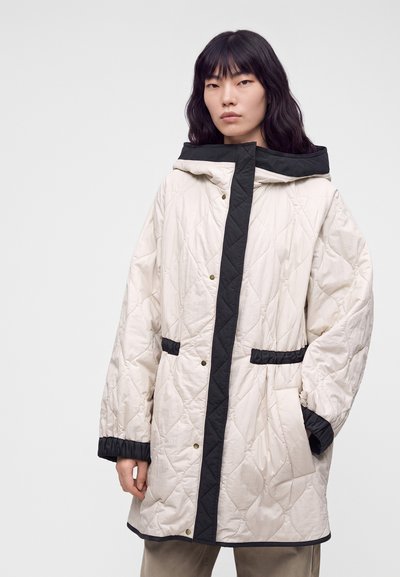 Femme portant un long manteau blanc matelassé avec des bordures noires et des poches, debout devant un fond clair uni.