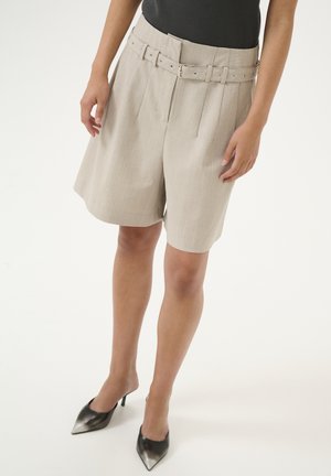 Beige high-waisted shorts made from textured fabric, featuring a belted waist, pleats, and a subtle check pattern, paired with black pointed heels.