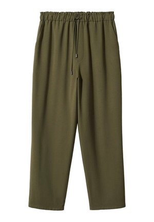 Kobieta ubrana w biały prążkowany sweter z podwiniętymi rękawami i oliwkowozielone spodnie ze sznurkiem, stoi z jedną ręką na biodrze, druga swobodna.