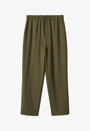 Kobieta ubrana w biały prążkowany sweter z podwiniętymi rękawami i oliwkowozielone spodnie ze sznurkiem, stoi z jedną ręką na biodrze, druga swobodna.