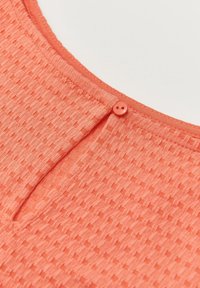 Tissu corail avec des motifs carrés texturés, doté d'un col arrondi et d'une petite fermeture à bouton assortie à l'arrière.