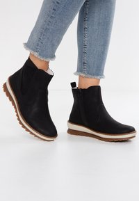 Svarta anklar stövlar gjorda av texturerat syntetmaterial, med sidodragkedjor, en vit sula och en brun gummisula.